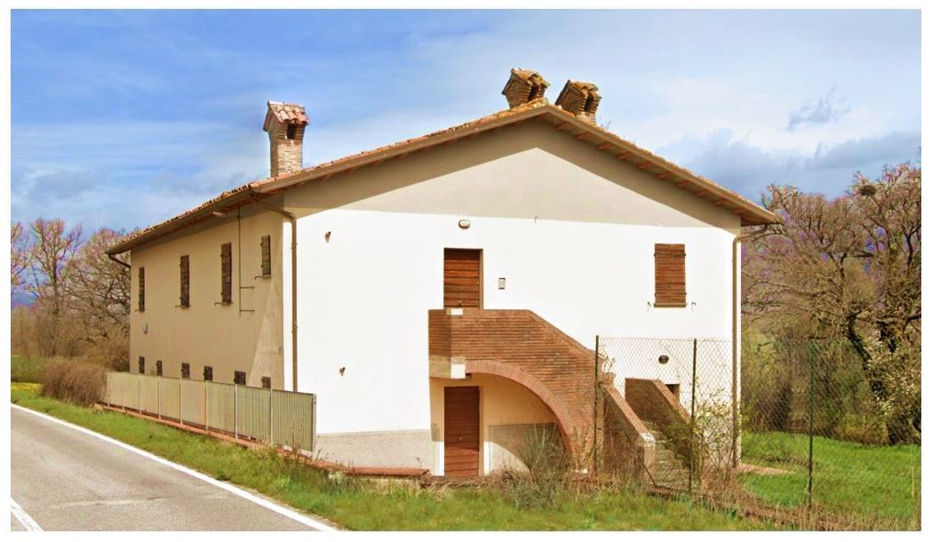 Casale di due piani con tetto spiovente a tegole su due falde, facciata beige chiaro con fasce decorative color tortora, infissi in legno naturale, scala ad arco rampante rivestita con mattoni rossi, porzione dalla strada provinciale 241 tra Gualdo Tadino a S.Pellegrino